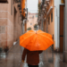 Girl with an orange umbrella protecting herself from the rain walking down a lonely narrow street