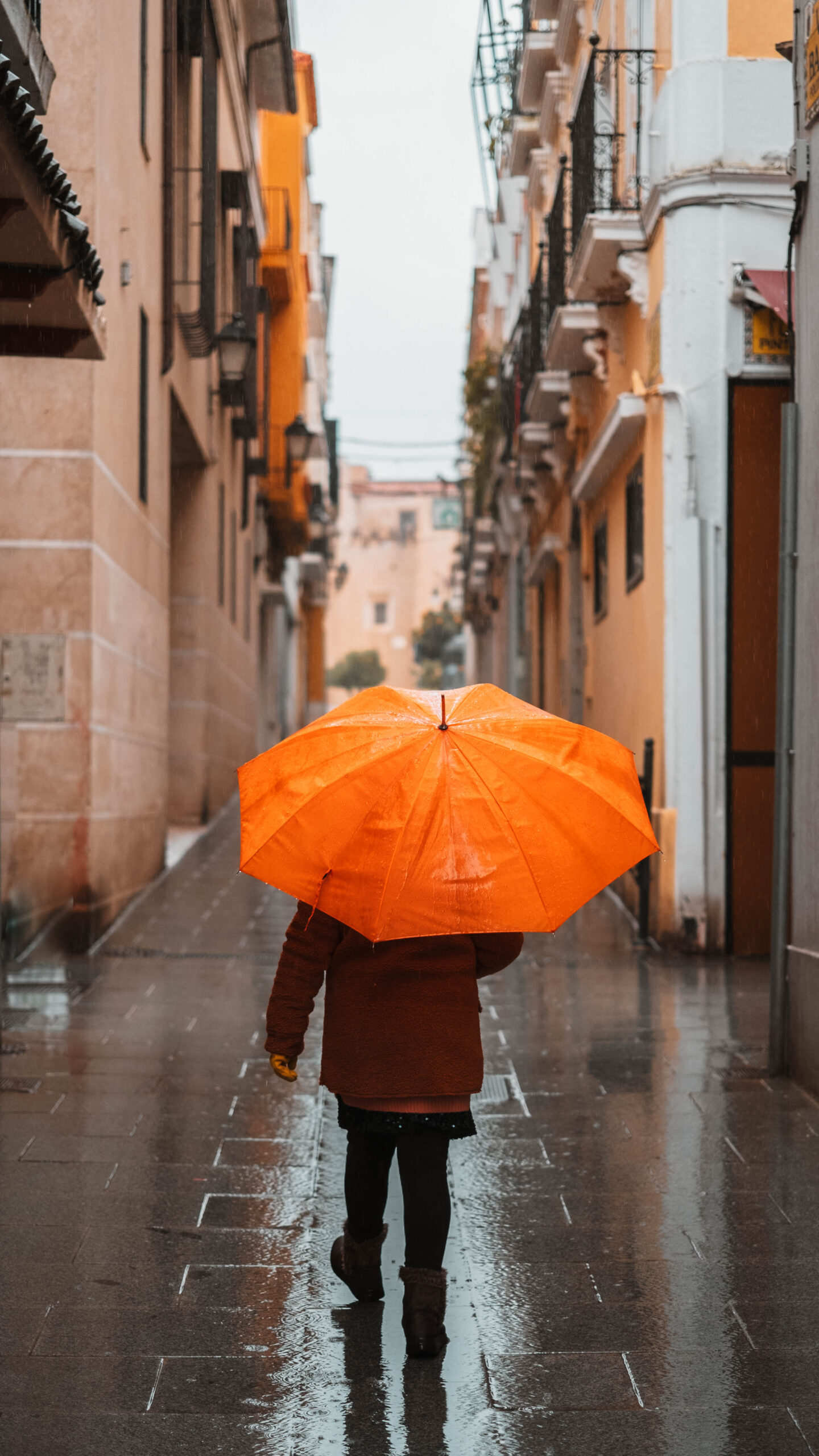 Girl with an orange umbrella protecting herself from the rain walking down a lonely narrow street